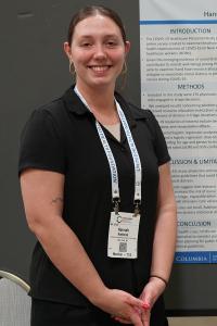 A smiling woman wearing a conference badge poses in front of her research poster